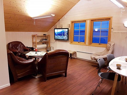 Gemütliches Wohnzimmer in Nord-Norwegen mit Ledersesseln, Holzmöbeln und Meerblick durch große Fenster. Ein Fernseher bietet Unterhaltung. Perfekt für Angelreisende, die Entspannung in einer komfortablen Umgebung suchen.