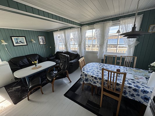 Gemütliches Wohnzimmer in Hitra, Norwegen, mit Holzwänden, Sofaecke, Esstisch mit geblümter Tischdecke und großen Fenstern mit Blick aufs Meer. Perfekt für einen entspannten Angelurlaub in malerischer Umgebung.