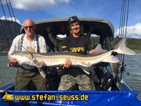 Zwei Männer halten einen großen Stör auf einem Boot, umgeben von Angelruten und unter einem blauen Himmel.