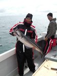 Eine Person in warmer Kleidung hält einen großen Fisch auf einem Boot, während eine weitere Person im Hintergrund angelt.