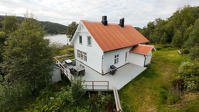 Einladendes weißes Ferienhaus in Norwegen mit rotem Dach, umgeben von grüner Natur. Große Terrasse mit Sitzmöglichkeiten, ideal für Angelreisen in Hitra. Malerischer Blick auf nahegelegenen See. Perfekt für Naturliebhaber und Ruhe suchende Reisende.