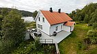 Einladendes weißes Ferienhaus in Norwegen mit rotem Dach, umgeben von grüner Natur. Große Terrasse mit Sitzmöglichkeiten, ideal für Angelreisen in Hitra. Malerischer Blick auf nahegelegenen See. Perfekt für Naturliebhaber und Ruhe suchende Reisende.
