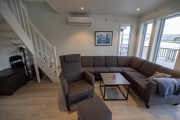 Gemütliches Ferienhaus auf Hitra, Norwegen. Modernes Wohnzimmer mit großen Fenstern, Ecksofa, Sessel und Treppe. Helle, einladende Atmosphäre mit schöner Aussicht auf die umliegende Natur. Perfekt für entspannte Angelurlaube.