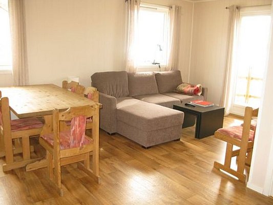 Gemütliches Wohnzimmer in Nord-Norwegen mit Holzboden, beiger Couch, Esstisch und Stühlen. Helle Einrichtung, ideal für einen entspannten Angelurlaub. Große Fenster lassen viel Tageslicht herein und schaffen eine einladende Atmosphäre.