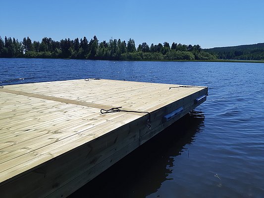 Ein hölzerner Steg schwimmt auf einem ruhigen See unter klarem, blauem Himmel, umgeben von Wald.