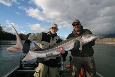 Zwei Personen halten einen großen Stör in einem Boot auf einem Fluss, umgeben von einer bewölkten Landschaft.