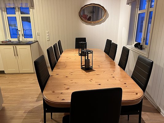 Gemütlicher Essbereich in Nord-Norwegen: Großer Holztisch, schwarze Stühle, Küchenzeile, Spiegel, rustikaler Stil. Ideal für geselliges Beisammensein nach abenteuerlichen Angelreisen im hohen Norden. Perfekter Ort für Erholung und Gemeinschaft.