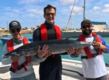 Drei Angler mit roter Schwimmweste präsentieren ihren beeindruckenden Fang, einen großen Fisch, auf einem Boot in der Küstenregion von São Vicente, Kapverden. Perfekter Angelurlaub für Abenteuer in exotischen Gewässern.