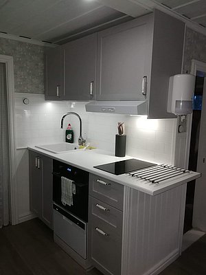Eine moderne Küche mit weißen Fliesen an der Wand, grauen Schränken, einem weißen Waschbecken, einem schwarzen Herd und einer dunkelgrauen Arbeitsplatte.