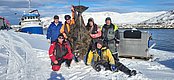 Sechs Angler präsentieren stolz ihren großen Fang, einen Heilbutt, im verschneiten Nord-Norwegen. Ein Fischkutter liegt im Hintergrund vor eisiger Küstenlandschaft vor Anker. Perfekte Bedingungen für dein nächstes Angelabenteuer.