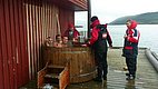 Fischer genießen eine Pause in einem hölzernen Outdoor-Whirlpool an einem Fjord in Nord-Norwegen. Die Szene zeigt eine entspannte Atmosphäre beim Angelurlaub in malerischer Umgebung nahe dem Wasser.
