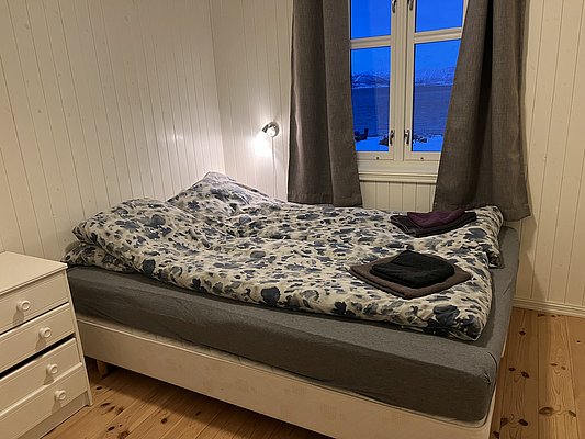 Gemütliches Schlafzimmer in Nord-Norwegen mit Doppelbett, Blumenbettwäsche und Fensterblick auf verschneite Landschaft. Ideal für erholsamen Angelurlaub in skandinavischer Natur. Rustikales Holzdesign ergänzt den Komfort für Angler auf der Suche nach Abenteuer.