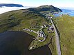Luftaufnahme einer malerischen Küstenlandschaft in Nord-Norwegen mit kleinen Häusern, Wohnmobilen, grünen Hügeln und dem offenen Meer im Hintergrund. Ideal für Angelurlaub und Naturabenteuer.