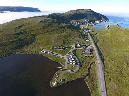 Luftaufnahme einer malerischen Küstenlandschaft in Nord-Norwegen mit kleinen Häusern, Wohnmobilen, grünen Hügeln und dem offenen Meer im Hintergrund. Ideal für Angelurlaub und Naturabenteuer.
