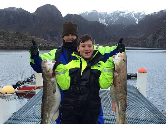 Zwei Angler auf einem Steg in Norwegen zeigen stolz ihren Fang: zwei große Kabeljau. Im Hintergrund beeindruckende Fjordlandschaft. Perfekter Angelurlaub in Hitra mit großartigen Fangmöglichkeiten und atemberaubender Natur.