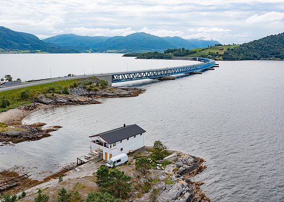 Küstenlandschaft von Hitra, Norwegen: Ein modernes Brückensystem überspannt das ruhige Wasser, umgeben von grünen Hügeln. Ein charmantes Haus steht nahe der Küste und verspricht Abenteuer und Erholung. Perfekt für Angelreisen.