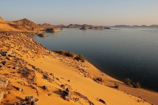 Eine sandige Wüstenlandschaft trifft auf einen weiten See mit felsigen Inseln, unter einem klaren Himmel in der Abenddämmerung.