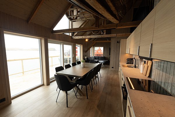 Modernes skandinavisches Innendesign in einer Hütte in Nordland, Norwegen. Geräumiges Esszimmer mit Tisch, großen Fenstern zum Meerblick und stilvoller Küche. Perfekt für Angelreisen in traumhafter Umgebung.