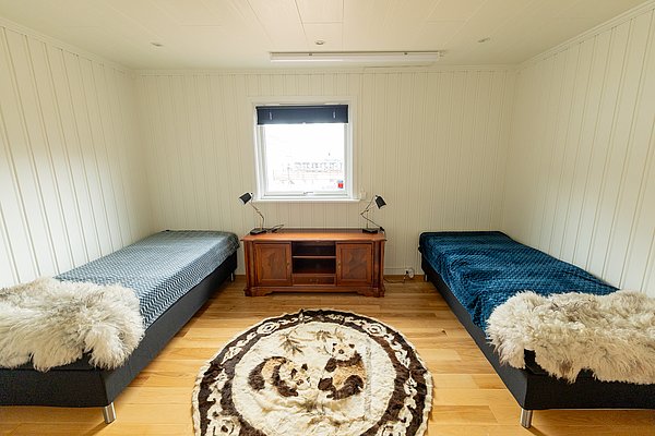 Gemütliches Zimmer in Nord-Norwegen mit zwei Einzelbetten, Holzboden und flauschigen Decken. Ein Fenster bietet Tageslicht, ein Teppich schmückt den Boden. Perfekt für entspannte Angelreisen in malerischer Umgebung.