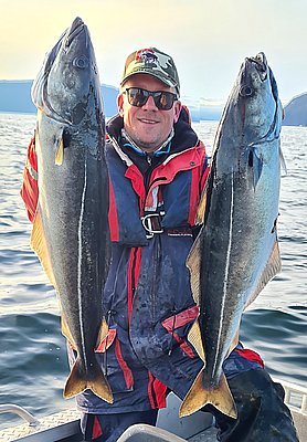 Ein Angler präsentiert zwei große Fische auf einem Boot in der Region Hitra, Norwegen. Die beeindruckende Küstenlandschaft im Hintergrund vermittelt Abenteuer und erfolgreiche Angelmöglichkeiten. Perfekt für Meeresangeln und Angelurlaub.