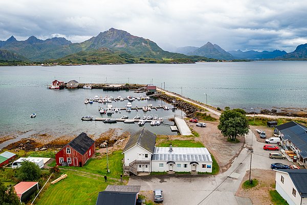 Luftaufnahme eines idyllischen Hafens in Nordland, Norwegen mit malerischen Bergen im Hintergrund. Kleine Boote liegen am Steg, umgeben von grüner Landschaft und typisch skandinavischen Häusern. Perfekt für Angelurlaub und Naturerlebnis.