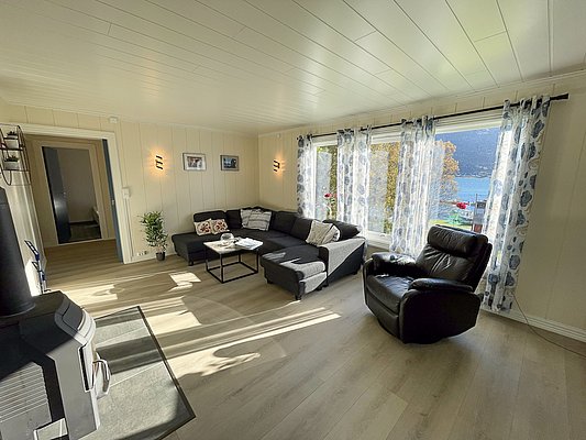 Helles Wohnzimmer in Nord-Norwegen mit gemütlicher Einrichtung, großem Ecksofa, Sessel und Kaminofen. Große Fenster bieten einen beeindruckenden Blick auf den Fjord. Perfekter Rückzugsort nach einem erfolgreichen Angelabenteuer im Norden.