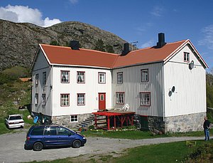 Großes, traditionelles weißes Haus mit roten Fensterrahmen und Dach, umgeben von grüner Natur und Bergen auf Hitra, Norwegen. Ideal für Angelurlaube mit atemberaubender Landschaft und Zugang zu hervorragenden Fischgründen. Perfekt für Erholung und Abenteuer.