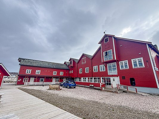 Rote Holzhäuser in Nord-Norwegen vor bewölktem Himmel. Traditionelle Architektur, ideal für Angelurlaube. Holzdielenweg, geparktes Auto, ruhige Atmosphäre. Perfekt für Abenteurer und Naturliebhaber. Entdecke die Schönheit der Region und genieße fischreiche Gewässer.