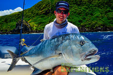 Ein begeisterter Angler präsentiert seinen beeindruckenden Fang, einen Giant Trevally, vor der traumhaften Küste Tansanias. Im Hintergrund sind dichter grüner Wald und tiefblaues Meer zu sehen. Perfektes Ziel für Angelreisen.