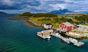 Küstenlandschaft in Nordland, Norwegen: Rotes Haus am Wasser, Anlegestelle mit Booten, grüne Inseln und Berge im Hintergrund unter bewölktem Himmel. Ideal für Angelurlaub und Naturerkundungen.