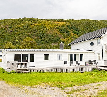 Weißes Haus am Fuße eines grünen Hügels in Nordland, Norwegen, ideal für Angelurlaube. Große Terrasse mit Blick auf die Natur, perfekt zum Entspannen nach einem erfolgreichen Angeltag.