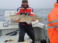Ein Angler in bunter Outdoor-Kleidung hält einen großen Kabeljau auf einem Boot im offenen Meer.