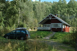 Rotes Holzhaus in Värmland, Schweden, umgeben von dichtem Wald. Ein Auto steht davor. Ideal für Angelurlaub und Naturerlebnisse in skandinavischer Landschaft. Friedliche Abgeschiedenheit und Abenteuer.