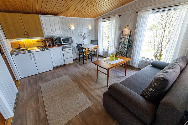 Gemütlicher Wohnbereich in einer norwegischen Hütte auf Hitra, Norwegen. Offene Küche, Esstisch, Sofa und große Fenster mit Ausblick auf die Natur. Perfekt für Angelreisende, die Komfort und Abenteuer vereinen möchten.
