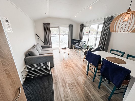 Gemütliches norwegisches Ferienhaus in Nord-Norwegen: Helle Einrichtung mit Holzfußboden, graue Couch, Esstisch und großem Fenster mit Fjordblick. Perfekte Ausgangsbasis für Angelerlebnisse und Entspannung in skandinavischem Ambiente.