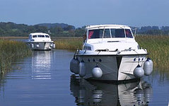 Zwei weiße Motorboote fahren auf einem ruhigen Fluss durch eine von Schilf gesäumte Landschaft.