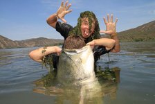 Ein Mann posiert spielerisch mit Pflanzen auf dem Kopf im Wasser und hält einen großen Fisch, während ein zweiter Mann im Hintergrund seine Zunge herausstreckt.