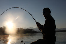 Ein Angler sitzt im Sonnenaufgang am Wasser und hält eine gebogene Angelrute.