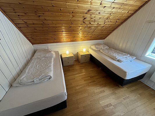 Gemütliches Schlafzimmer in Hitra, Norwegen, mit zwei Einzelbetten unter schräge Holzdecke. Weiße Wände, Holzboden und Nachttisch mit Lampen sorgen für eine einladende Atmosphäre. Ideal für Ruhesuchende nach einem Angeltag.