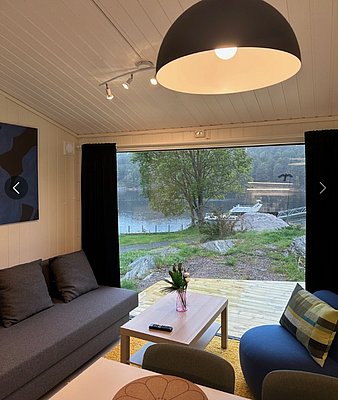 Gemütliches Wohnzimmer in Hitra, Norwegen, mit Fenstern zur Seelandschaft. Moderne Einrichtung, graues Sofa, Holztisch, sanfte Beleuchtung. Ideal für Angelreisen und naturverbundene Erholung. Perfekte Kulisse für Entspannung und Abenteuer gleichermaßen.
