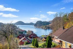 Küstenlandschaft in Süd-Norwegen: Malerische Holzhäuser vor einem ruhigen Fjord mit bewaldeten Hügeln unter strahlend blauem Himmel – ideales Reiseziel für entspannte Angelurlaube und Naturentdeckungen.