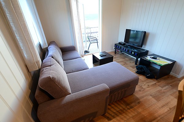 Gemütliches Wohnzimmer in Nord-Norwegen mit L-förmigem Sofa, Fernseher und Zugang zu einem Balkon mit beeindruckendem Ausblick. Perfekt für erholsame Angelreisen.
