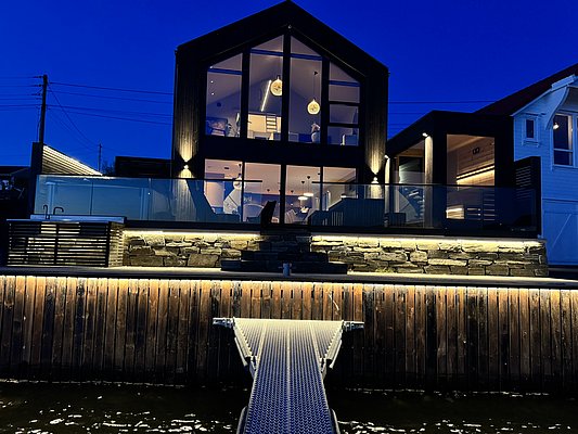 Modernes Haus am Wasser in Südwest-Norwegen, stimmungsvoll beleuchtet bei Nacht. Große Fensterfronten bieten Einblicke ins stilvolle Interieur. Perfekter Ausgangspunkt für Angelurlaube in Norwegen. Entdecke Abenteuer und Komfort in skandinavischer Idylle.
