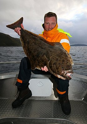 Eine Person hält einen beeindruckenden, großen Heilbutt auf einem Boot in Nord-Norwegen. Der Hintergrund zeigt die raue, wunderschöne Küstenlandschaft unter einem bewölkten Himmel. Perfektes Ziel für Meeresangeln-Enthusiasten.