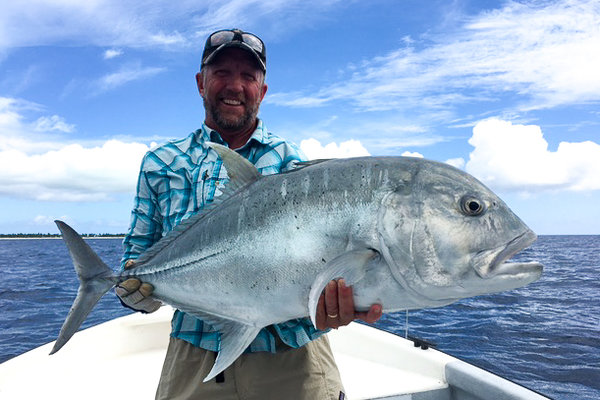 Ein Angler hält einen großen Giant Trevally auf einem Boot vor der Küste von Kiritimati, Kiribati. Blauer Himmel und ruhiges Meer im Hintergrund. Ideal für Angelreisen und Meeresangeln-Abenteuer.