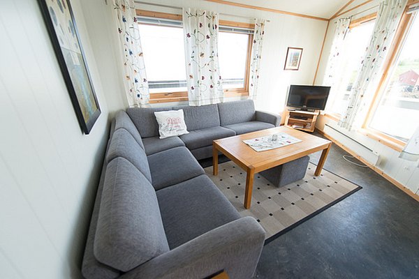 Gemütliches Wohnzimmer in Nordland, Norwegen. Helle, einladende Einrichtung mit großer grauer Eckcouch, Holztisch, Fernseher und dekorativen Vorhängen. Perfekt für entspannte Angelreisen und gemütliche Abende nach einem langen Tag am Wasser.