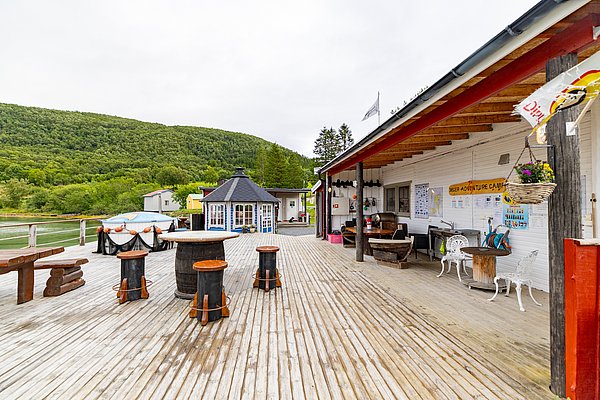 Angelcamp in Nord-Norwegen mit rustikaler Terrasse, Fässern und Sitzgelegenheiten, umgeben von grünen Wäldern. Ideal für entspannte Angelurlaube und Erkundungen in der unberührten Natur. Perfekter Ausgangspunkt für Abenteuer und Meeresangeln.