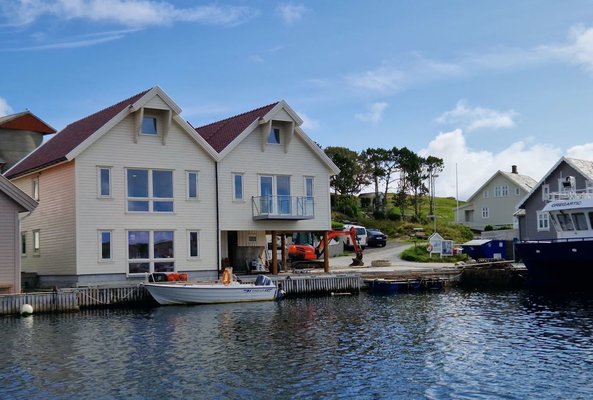 Zwei moderne Holzhäuser mit roten Dächern stehen am Ufer einer ruhigen Bucht, neben einem kleinen Boot und einem größeren blauen Schiff unter blauem Himmel. Zwei moderne Holzhäuser mit roten Dächern stehen am Ufer einer ruhigen Bucht, neben einem kleinen Boot und einem größeren blauen Schiff unter blauem Himmel.