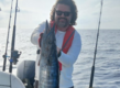 Ein Angler hält auf seinem Boot stolz einen großen Fisch vor der Küste von São Vicente, Kapverden. Der klare Himmel und das ruhige Meer unterstreichen das perfekte Angelabenteuer.