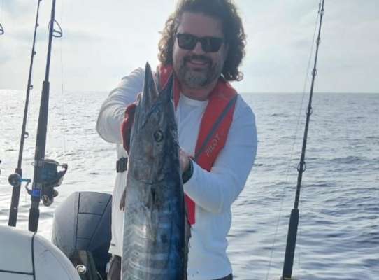 Ein Angler hält auf seinem Boot stolz einen großen Fisch vor der Küste von São Vicente, Kapverden. Der klare Himmel und das ruhige Meer unterstreichen das perfekte Angelabenteuer.